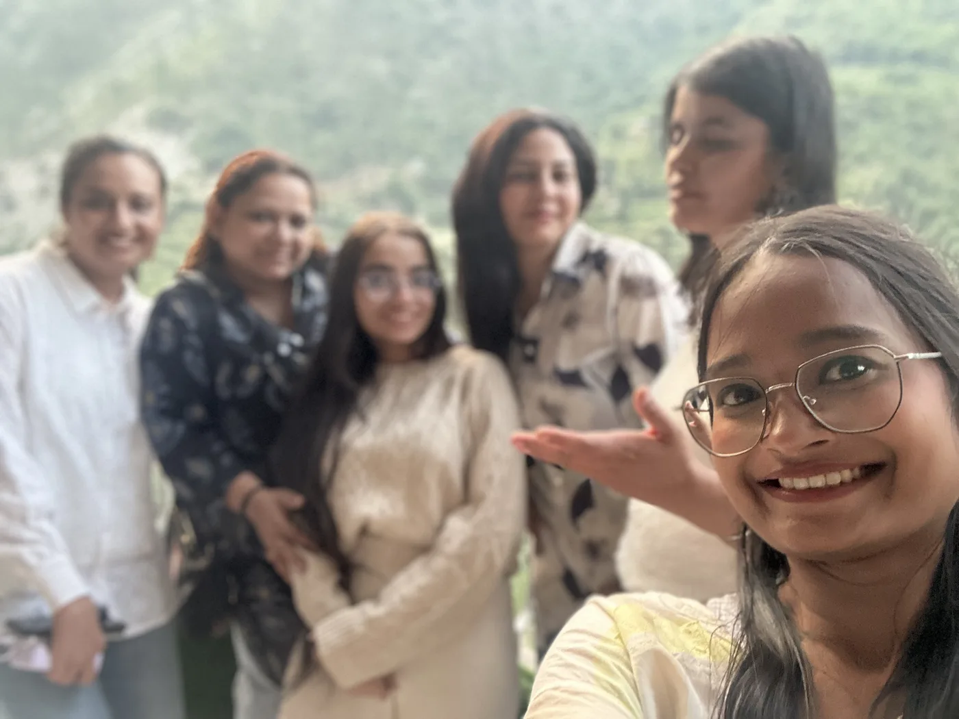 Six women crowded into a selfie on a hillside at dusk, the woman holding the camera laughing in the foreground and green ridges behind.