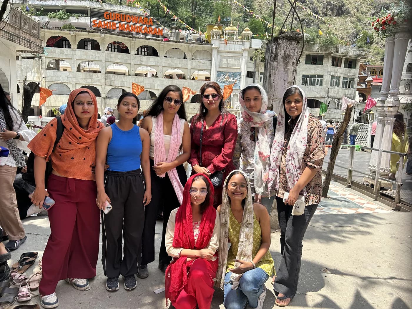 Eight women standing together on the marble forecourt of Gurudwara Sahib Manikaran, heads covered, with the gurudwara and a steep valley wall behind them.