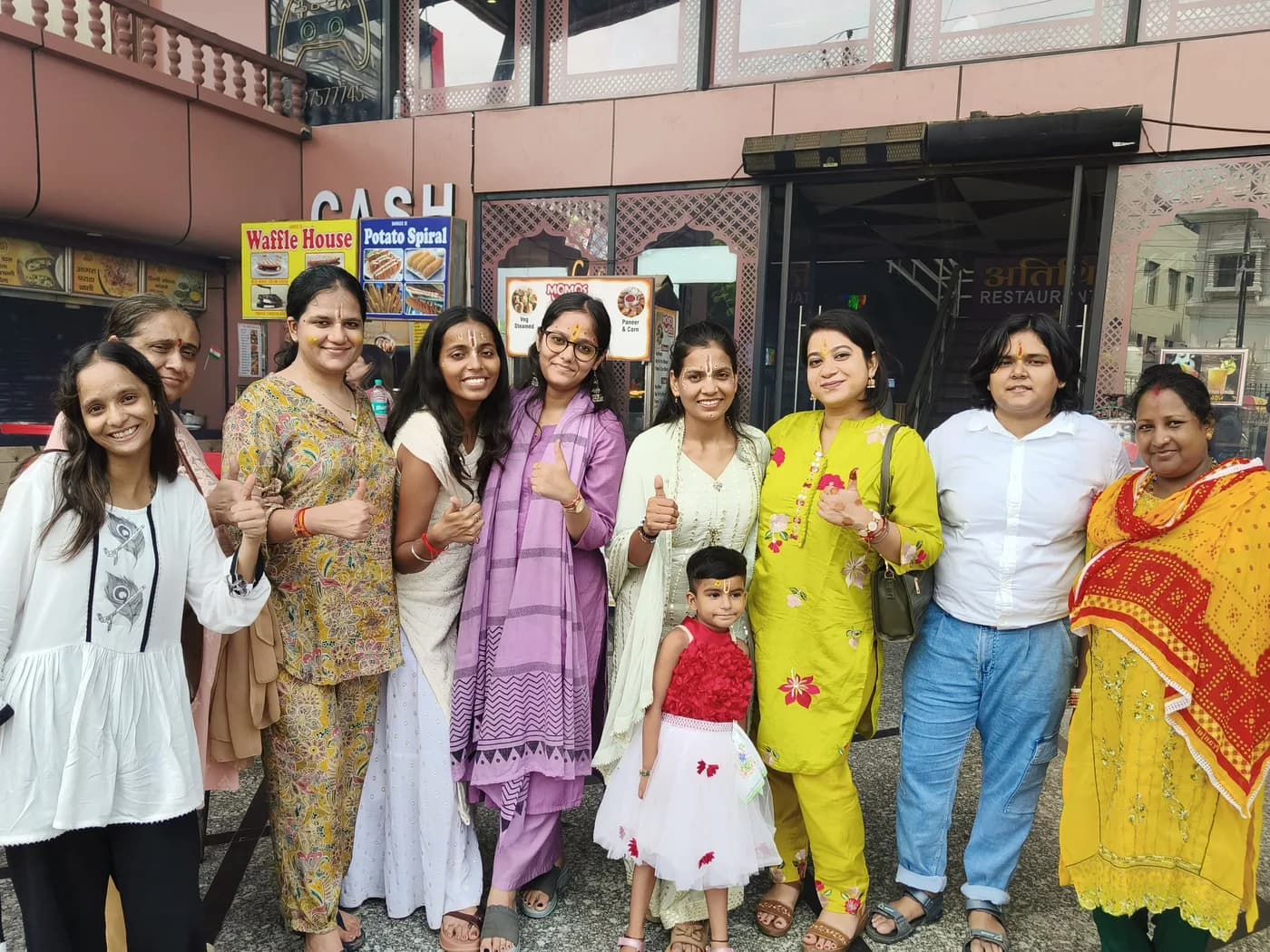 Ten women and a small girl standing together outside a row of street-food counters, most giving a thumbs-up to the camera.
