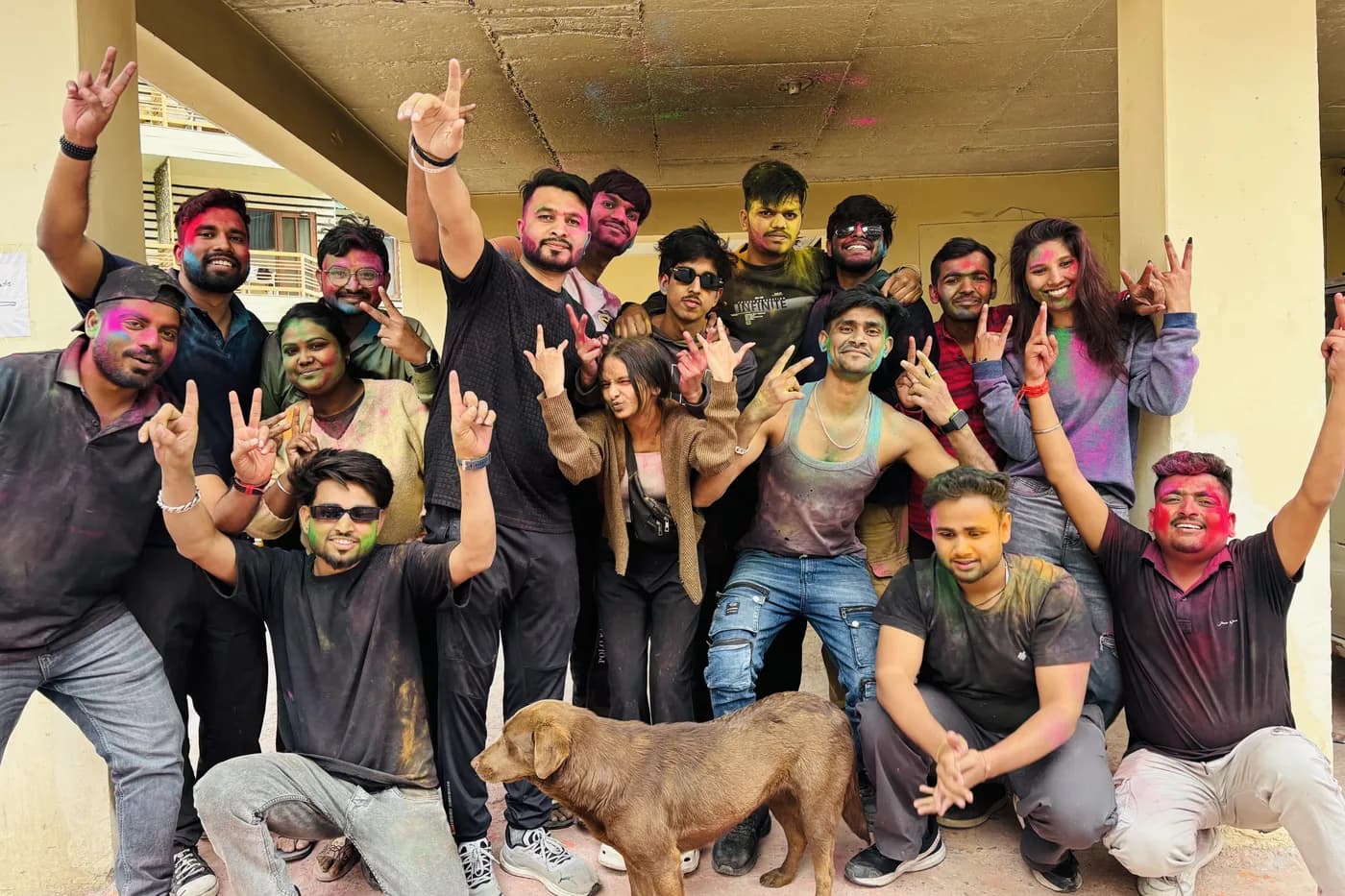 A large group covered head to toe in Holi colours, arms up, cheering in a concrete courtyard, with a street dog wandering through the middle of the shot.
