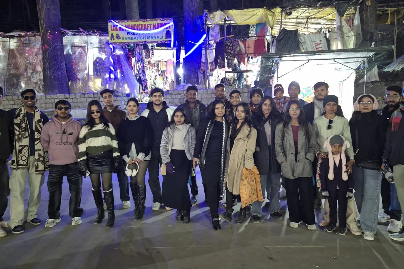 A group of about twenty-two in winter coats standing in a lit handicraft market at night, stalls strung with lights behind them.