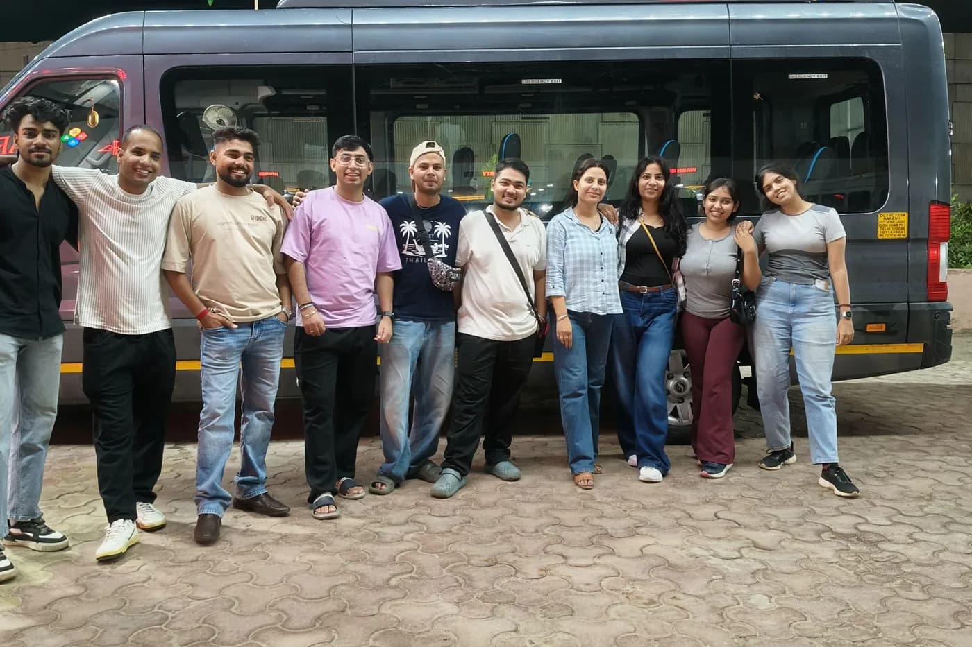 Eleven travellers standing shoulder to shoulder in front of a tempo traveller minibus at night, luggage stowed.